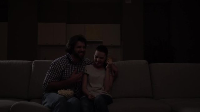 Couple sits on sofa and wathes movie. Guy has bowl with popcorn on knees. They start to laugh. People are watching comedy. Girl is pointing forward and looking at guy.