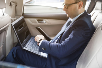 a young business man in a suit is sitting in the back seat of a business car with a laptop and a notebook, business negotiations