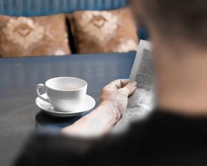 A man reading newspaper and drinking coffee sitting in cafe or at home in kitchen at morning time. A coffee cup on dark blue table. A person hands holding newspaper. Back and nape are blurred