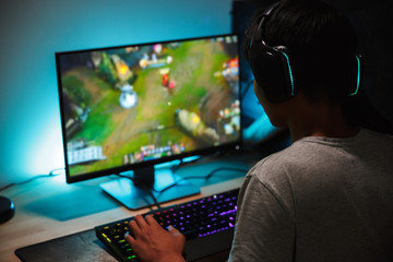 Image of immersed teenage gamer boy playing video games on computer in dark room, wearing headphones and using backlit colorful keyboard