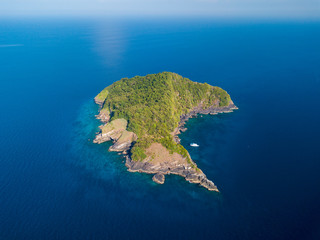 A single boat next to a remote, uninhabited tropical island covered in green jungle and surrounded by tropical coral reef