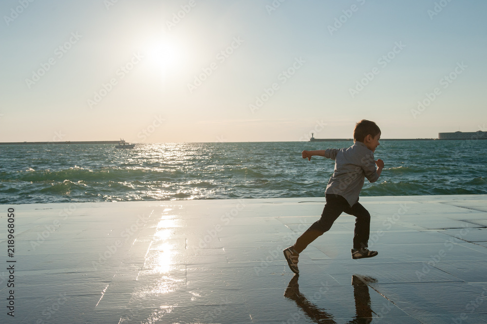 Wall mural healthy caucasian kid running along sea coast in port city in autumn sunset