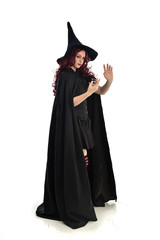 full length portrait of red haired girl wearing long black cloak, pointy hat and witch costume. standing pose, isolated on white studio background.