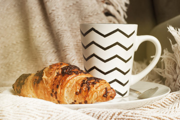 Cup of coffee with croissant on a white plate, with warm blanket. Breakfast, selective focus, toned.