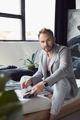 Handsome man with dark hair and beard in light jacket and linen pants sits on the bed in scandinavian studio with big window and plants