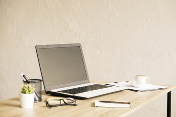 Blank screen laptop computer, cup of cappuccino coffee, cactus, supplies and folded eye glasses on wooden desk in spacious office full of sunlight. Creative workspace. Close up, copy space, background