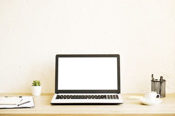 Fototapeta premium Blank screen laptop computer, cup of cappuccino coffee, cactus, supplies and folded eye glasses on wooden desk in spacious office full of sunlight. Creative workspace. Close up, copy space, background