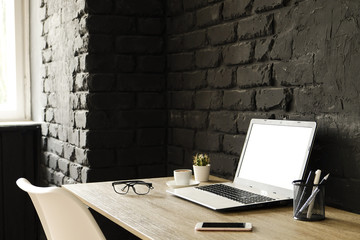 Creative workspace of a blogger. White laptop computer & folded eye glasses on wooden table in loft style office with black brick walls. Designer's table concept. Close up, copy space, background.