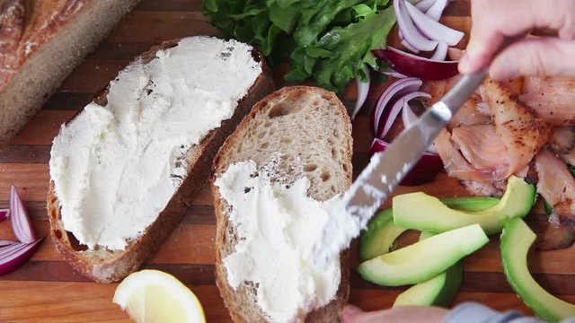A woman adds mayonnaise, smoked salmon slices, avocado, onion and lettuce to artisan bread slices