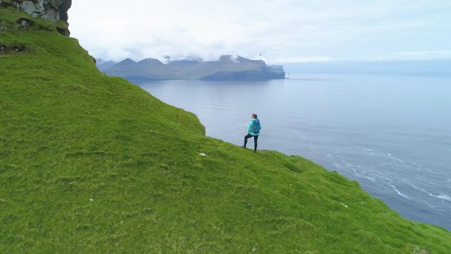 AERIAL: Flying above woman photographing the picturesque Scandinavian seaside from the edge of a cliff on grassy mountainous island in Faroe Islands. Unrecognizable photographer taking photos of ocean