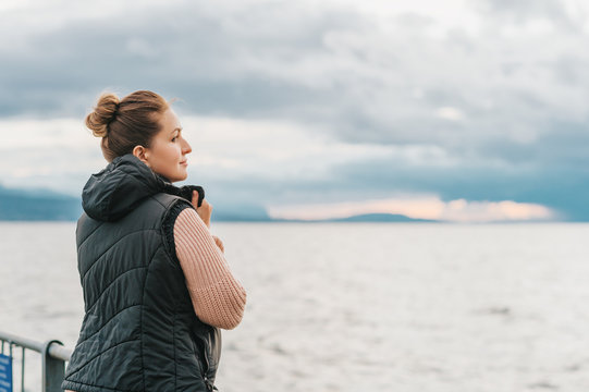 Outdoor portrait of young woman resting by the lake on a fresh cold day, wearing warm pullover and black down vest