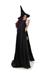 full length portrait of red haired girl wearing long black cloak, pointy hat and witch costume. standing pose, isolated on white studio background.