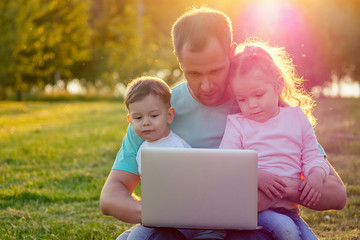 father and his cute two baby little girl and boy sitting on grass looking at laptop in summer park