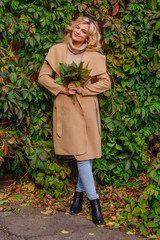 Young beautiful woman in sweater and coat stands next to the background of wild grapes holding bouquet of autumn leaves