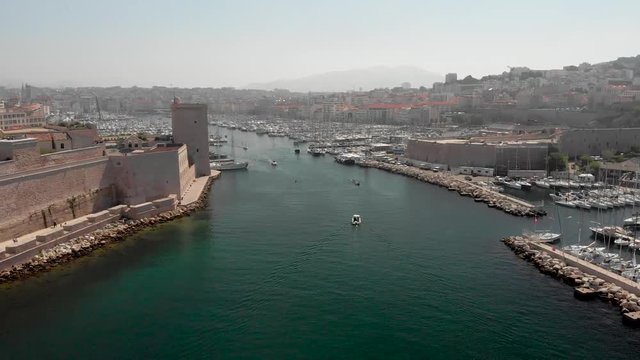 Flying into the old port of Marseille in aerial drone shot. French marina with yachts and boats traveling by historic castle and phare de sainte marie lighthouse into cafetiere. 