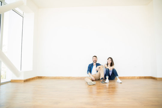 Smiling Couple Sitting In Empty Room Of New Apartment
