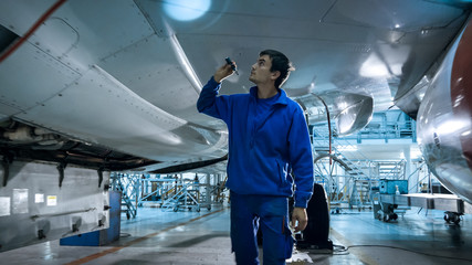 Aircraft maintenance mechanic with a flash light inspects plane fuselage in a hangar.