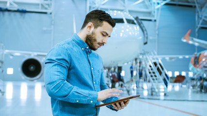 In Big Company Hangar Aircraft Maintenance Engineer Uses Tablet Computer while Standing Near Big New Shiny White Plane.