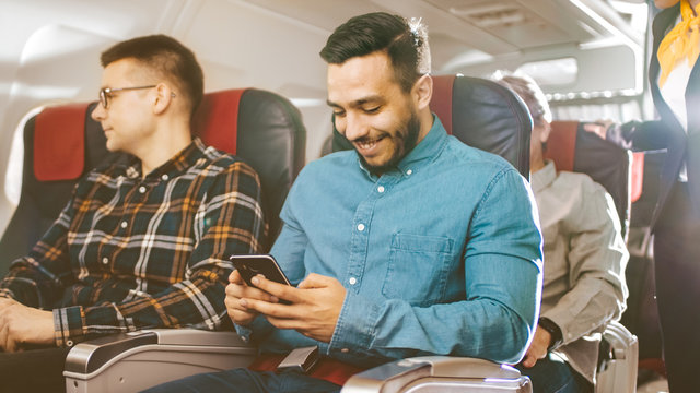 On a Commercial Flight Young Hispanic Male Uses Smartphone, while His Neighbor Looks out of the Window. Senior Man in the Back Sleeps Peacefully.