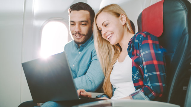 On a Board of Commercial Airplane Beautiful Young Blonde with Handsome Hispanic Male Watch Movies on a Laptop and Laugh. Sun Shines Through Aeroplane Window.