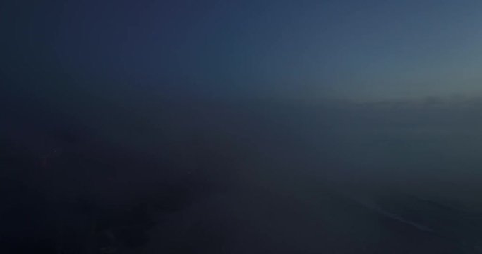 Aerial shot through fog clod to reveal cliff overlooking ocean and beach at blue hour. Then dollies back into fog.