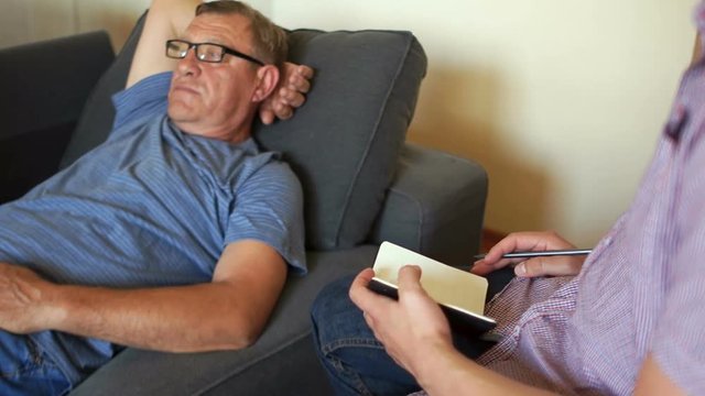 Conversation with a psychologist. The pensioner talks to the psychiatrist lying on the couch. The man takes off his glasses and rubs his eyes. The psychologist makes notes in the notebook