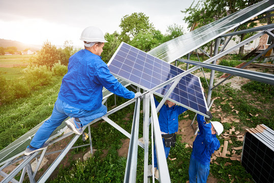 Installing of stand-alone solar photo voltaic panel system. Team of three technicians in hard-hats mounting the solar module on platform on green summer view background. Alternative energy concept.