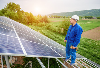 Construction worker with screwdriver standing on metal frame of photo voltaic solar system looking on shiny surface. Alternative energy, ecology protection and cheap electricity production concept.