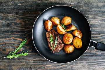 Fried potatoes with beef steak with rosemary in a frying pan on a wooden table