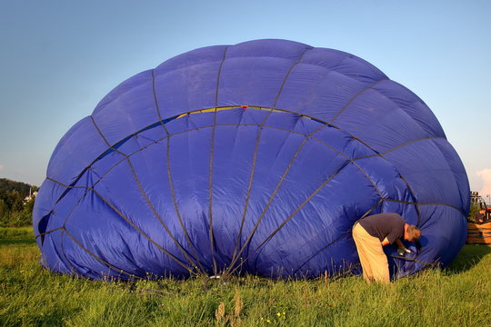 Na wpół napopmpowany powietrzem wielki niebieski balon leży na trawiastym podłożu, członek załogi dociąga ostatnie sznury w czaszy balonu, widok z tyłu, niebieskie niebo w tle