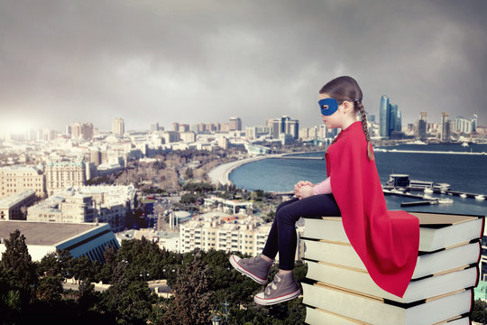 Superhero kid sitting on a stack of books against urban background. Girl power concept