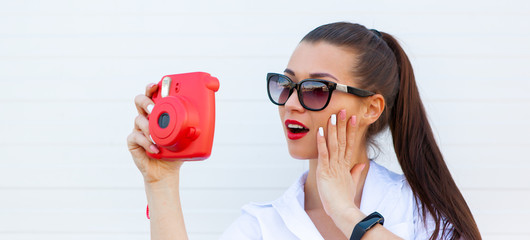 Fashion portrait of pretty smiling woman in sunglasses making photo by the camera against the grey wall. Wireless Headphones, fitness bracelet on the hand