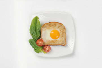 Slice of fried bread with egg on a white plate. On a plate next to the toast, fresh spinach greens and cherry tomatoes. White background. Close-up. View from above.