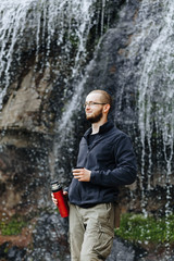 Man drinks tea or coffee from a thermos, standing near a waterfall in the mountains