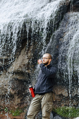 Fototapeta na wymiar Man drinks tea or coffee from a thermos, standing near a waterfall in the mountains