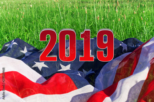 The background of the American flag and 2019. The flag of the USA lies on the green grass, and on it the voluminous figures are 2019.