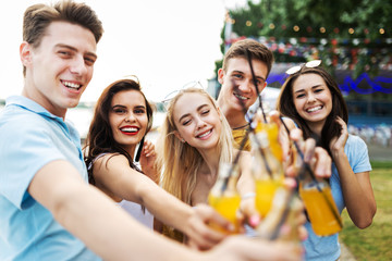 A company of good-looking friends laughing and drinking yellow cocktails and socialising  in the nice cafe next to the river. Cheers. Entertainment, having good time. River is in the background.