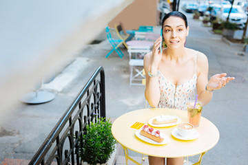 Emotional talk. Emotional young woman having an important phone talk while sitting outdoors at the table covered with desserts and drinks