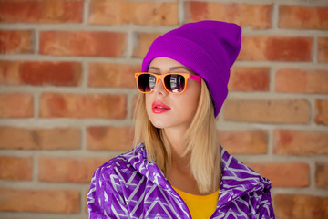 Young blonde girl in 90s sports jacket and hat on brick wall background