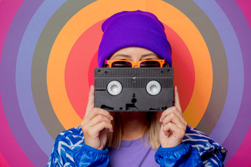 Young blonde girl in 90s sports jacket and hat with cassette on rainbow background