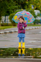 Happy funny child with multicolored umbrella jumping puddles in rubber boots and laughing