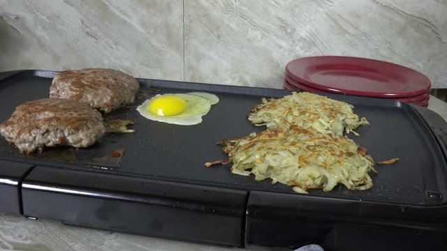 Frying an egg on an electric grill with sausage and hash browns