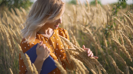 Beautiful happy young woman in field of spikelets and wheat with book on the sunset, blonde in the grass having a good time