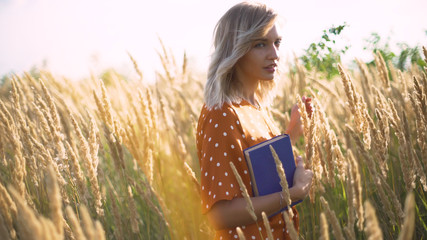 Beautiful happy young woman in field of spikelets and wheat with book on the sunset, blonde in the grass having a good time