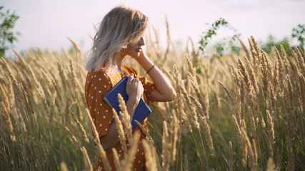 Beautiful happy young woman in field of spikelets and wheat with book on the sunset, blonde in the...