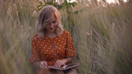 Beautiful happy young woman in field of spikelets and wheat with book on the sunset, blonde in the...
