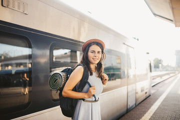 theme railway and travel. Portrait young caucasian woman with toothy smile standing at train station train background with backpack and equipment for tourism isolated mat in dress and hat in summer