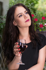 Young beautiful girl holding glass with red wine and relaxing in garden park on sunny summer day, woman is in black elegant dress. Close up, selective focus