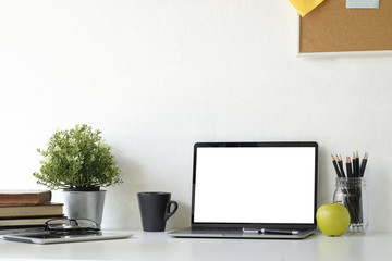 Mockup laptop computer on Stylish table Board and post it, book, pencil, glasses  and coffee mug with tablet on table with wall.