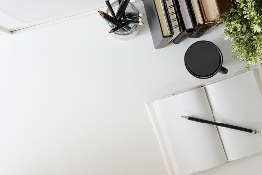 Flat lay, top view office table desk. Workspace with notebook, book, office supplies, pencil, green leaf, and coffee cup on white desk.